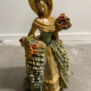 Dekorativ figur av kvinna med blommor - Charmig dekorativ figur föreställande en kvinna i klänning med blommor i händerna. Figuren har en mjuk färgpalett med inslag av grönt, gult och rött. Perfekt som prydnad i hemmet.