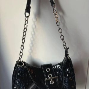 Svart axelväska med kedjedetaljer - Snygg svart axelväska i syntet med glansig finish och kedjedetaljer. Väskan har en dragkedja och ett bälte med spänne framtill. Perfekt för att ge din outfit en edgy touch.