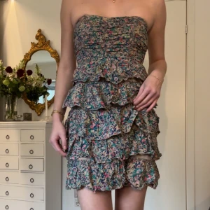 Blommig axelbandslös klänning - Storlek s! Säljer en söt axelbandslös klänning med blommigt mönster och volanger. Perfekt för sommarens fester. Klänningen har en kort längd och en härlig passform.