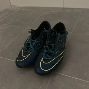 Nike Mercurial fotbollsskor - Snygga Nike Mercurial fotbollsskor i blått med gula detaljer och stänkmönster. Skorna har snörning och är perfekta för fotbollsplanen. De har en smidig och tight passform för optimal kontroll och komfort. OBS! Skruvdobbar!