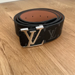 Louis Vuitton svart skärp med LV-spänne - Svart Louis Vuitton-bälte i skinn med klassiskt LV-monogram och stor silvrig LV-logga som spänne. Insidan är ljusbrun och bältet har präglad märkning. Perfekt accessoar för att lyfta din outfit med lyxig känsla.