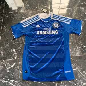 Chelsea David Luiz 4 Adidas tröja - Chelsea FC fotbollströja från Adidas med David Luiz och nummer 4 på ryggen. Blå med vita detaljer, klassiska Adidas-ränder på axlarna och Samsung som sponsor. Tillverkad i lätt och ventilerande ClimaCool-material, perfekt för match eller träning.