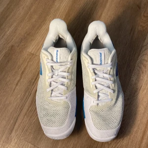 Vita och blå Babolat tennisskor - Snygga tennisskor från Babolat i vitt och blått med mesh-ovandel för bra ventilation. Skorna har snörning, platt sula och förstärkta partier för extra stabilitet. Perfekta för dig som vill ha stilrena och sportiga skor på tennisbanan.
