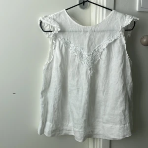 Vit ärmlös blus med spets från Zara - Supersöt vit blus från Zara i luftig linnemix. Ärmlös modell med volangdetaljer vid axlarna och spetsdekor framtill. Blusen har v-ringning. Knappen som man stänger med har gått av (som man kan se på bilden) men det är bara att sy på en ny. Pris går att diskutera 