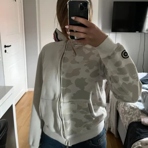 BAPE camo hoodie med hajhuva, beige - Säljer en beige hoodie från BAPE med ikoniskt camo-mönster och hajdetalj på huvan. Tröjan har dragkedja, känguruficka och BAPE-logga på ärmen. Perfekt för dig som gillar streetwear och vill sticka ut med en unik look.
