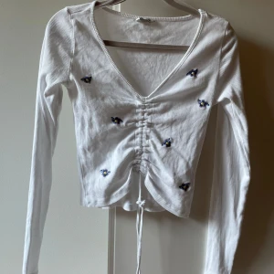 Vit somrig blus med broderade blommor - Supersöt vit croppad blus från Cubus med broderade små blommor i lila. Toppen har lång ärm, v-ringning och snörning framtill som ger en snygg rynkad effekt. Ribbad bomullskvalitet som känns mjuk mot huden. Använd 2-3 gånger så ser ny ut.