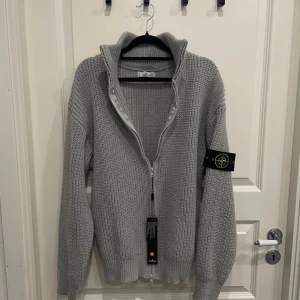 Stone Island Cardigan - Säljer en snygg ljusgrå stickad kofta från Stone Island med dragkedja framtill och hög krage. Klassisk patch på ärmen och ribbade muddar. Perfekt för lager-på-lager och stilren look.