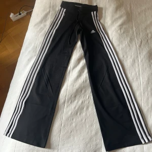 Svarta Adidas byxor med vita ränder - Svarta Adidas byxor med klassiska tre vita ränder längs benen och Adidas-logga på låret. Byxorna har snörning i midjan och är raka i modellen. 