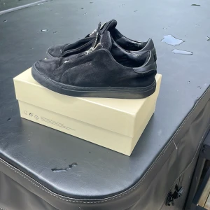 Svarta sneakers från Axel Arigato mocka - Tjenare! Jag säljer mina Axel Arigato skor. Dem är använda men har kvar att ge. Det finns en  logga på sidan där det står Arigato men tyvärr har loggan försvunnit på ena skon. På undersulorna har det fastnat små löv som jag ej fick bort. På höger skon har en söm släppt, men inget som påverkar skon. De kommer med en box och en påse som är original.