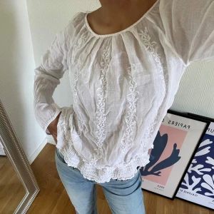 Vit blus - Superfin vit blus från vero Moda med broderade detaljer framtill och smockad nederkant. Lätt och luftig i bomull, perfekt till jeans eller kjol. Långärmad med rund halsringning och romantisk vibe. 