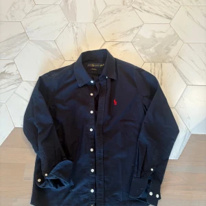 Mörkblå skjorta från Ralph Lauren - Klassisk mörkblå skjorta från Ralph Lauren med vit knappstängning och röd broderad logga på bröstet. Skjortan har lång ärm, normal passform och är tillverkad i bomull. Perfekt för en stilren och tidlös look. Perfekt för dig som vill ha en fräsch skjorta för sommar kvällar! Priser är ej hugget i sten, hör av er!