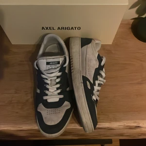 Axel Arigato sneakers i vitt och marinblått - Snygga sneakers från Axel Arigato i vitt och marinblått. Skorna har klassisk låg siluett, rund tå och snörning. Ovandelen är i skinn med perforerade detaljer på tån och sidorna. Sulan är vit med tydlig Arigato-logga undertill. Perfekta för en clean och modern stil. Modellen säljs inte längre.