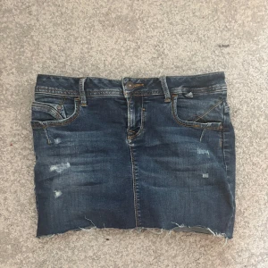  Lågmidjad jeanskjol  från LTB - Snygg mörkblå jeanskjol från LTB med slitna detaljer och rå kant nertill. Klassisk femficksmodell med synliga sömmar i kontrastfärg och bälteshällor. Mycket bra kvalité.