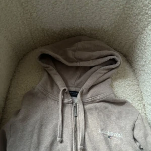 Beige hoodie med dragkedja från Lexington - Mysig beige hoodie från Lexington med dragkedja och huva. Tröjan har snörning vid huvan och broderad Lexington-logga på bröstet. Perfekt för chill dagar och enkel att matcha med jeans eller mjukisbyxor.