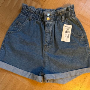 Högmidjade jeansshorts KappAhl 164 - Snygga blå jeansshorts från KappAhl med hög midja och elastisk, rynkad linning. Shortsen har två framfickor, två bakfickor och uppvikta benslut. Stängs med två färgade knappar och dragkedja. Perfekta för sommaren och riktigt sköna att bära.