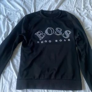 Svart sweatshirt från Hugo Boss - Svart sweatshirt från Hugo Boss med stor vit broderad logga på bröstet. Tröjan har rund hals, långa ärmar och ribbade muddar. Snygg och stilren modell som passar perfekt till jeans eller joggers.