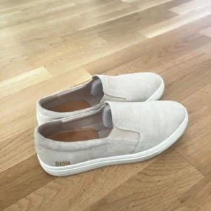 Beige slip-on sneakers från Dasia/Novita - Snygga slip-on sneakers i beige mocka från Dasia och Novita. Skorna har vit platt sula och rund tå, perfekt för en clean och stilren look. Lätta att ta på och av tack vare elastiska partier på sidorna. Passar till allt och ger en fräsch vibe. Använt ett fåtalgånger( pris kan diskuteras) 