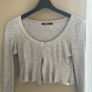Grå croppad topp med spets från Gina Tricot - Supersöt ljusgrå croppad topp från Gina Tricot med långa ärmar, spetskant runt halsringningen och framtill samt en liten rosett. Toppens material är mjukt och stretchigt, perfekt för en chill och trendig look.