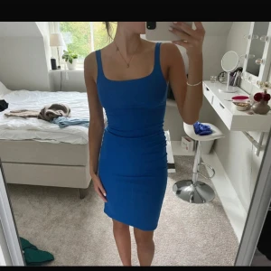 Kornblå tight kortklänning - Säljer en kornblå tight kortklänning med breda axelband och fyrkantig urringning. Klänningen har en figurnära passform och en liten slits bak för extra rörelsefrihet. Perfekt för dig som gillar en enkel men snygg look.