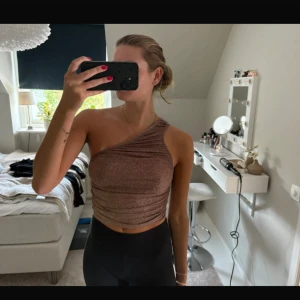 Glittrig brun one shoulder topp - Säljer en glittrig brun croppad topp med one shoulder-design. Toppen har en tight passform och är tillverkad i ett stretchigt syntetmaterial som ger en snygg glansig effekt. Perfekt för dig som vill sticka ut med en trendig och cool look.