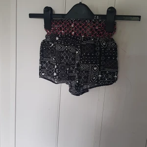 Svarta mönstrade shorts Revival - Snygga svarta shorts från Revival med vitt grafiskt mönster och hjärtan. De har en bred, smockad midja i svart och rosa som ger en cool kontrast. Lätta och luftiga, perfekta för varma dagar. Passformen är loose och midjan är hög.