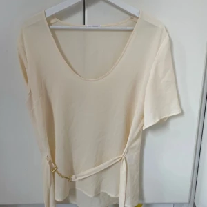 Beige omlottopp med knyt och gulddetalj - Snygg beige omlottopp med kort ärm och djup rundringning. Toppen har ett knytband i midjan med guldfärgad metallspänne som ger en extra detalj. Perfekt för dig som gillar stilrena och trendiga plagg.
