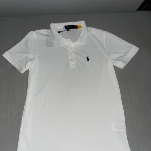 Vit pikétröja från Polo Ralph Lauren - Klassisk vit pikétröja från Polo Ralph Lauren med korta ärmar och broderad logga på bröstet. Tröjan har krage och knäppning framtill. Perfekt för en clean och stilren look. Tillverkad i bomull för en skön känsla.