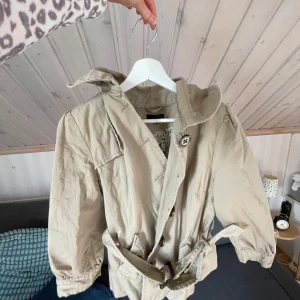 Beige trenchcoat från H&M med bälte - Snygg beige trenchcoat från H&M i storlek 40. Jackan har knappar framtill, bälte i midjan och trekvartsärmar. Insidan är fodrad med blommigt tyg i grönt och brunt. Perfekt för vår och höst när du vill ha en chill men stilren look.