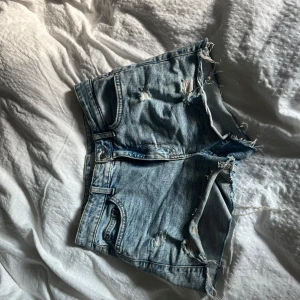 Blå slitna jeansshorts från Cubus - Säljer ett par blå jeansshorts från C Denim med råa, fransiga kanter och slitna detaljer. Shortsen har klassisk femficksdesign, bälteshällor och knappstängning. Perfekta för en avslappnad och edgy stil i sommar.
