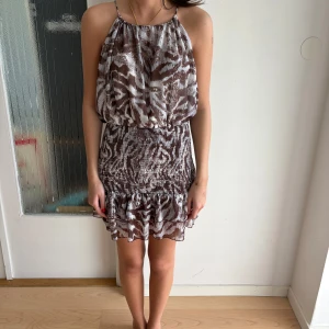 Brun zebramönstrad kortklänning Zara - Supersöt kort klänning från Zara i brunvitt zebramönster. Den har smala axelband, volang nertill och smockad midja som ger en snygg passform. Tunt, luftigt material som känns perfekt för varma dagar. Perfekt för dig som gillar trendiga mönster och en avslappnad men chic stil.