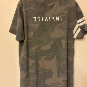 Grå camo t-shirt River Island M - Snygg grå camo t-shirt från River Island i storlek M. T-shirten har korta ärmar med vita ränder på ena ärmen och texten 'INFINITE' tryckt på ryggen. Perfekt för dig som gillar streetstyle och vill sticka ut lite extra.