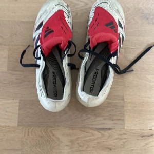 Adidas Predator fotbollsskor röd/vit tounge  använt en gång och är i jättebra skick  - Säljer ett par Adidas Predator fotbollsskor i vitt med röda och svarta detaljer. Skorna har snörning, täckt snörparti och dobbar för gräsplan. Ovandelen är i syntetmaterial med Hyperpeel-textur för extra grepp på bollen. Perfekta för dig som vill sticka ut på planen.