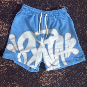 Blåa Syna shorts - Blå shorts med stort vitt graffiti-inspirerat tryck framtill och coola snören i midjan. De har elastisk midja och är perfekta för en avslappnad streetwear-look. Materialet känns mjukt och lätt, perfekt för varma dagar.