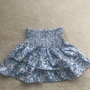 Blåvit volangkjol från Gina Tricot - Supersöt kort volangkjol från Gina Tricot i blått och vitt med mönster. Kjolen har smock i midjan och dubbla volanger som ger en flowy känsla. Perfekt för sommarens alla dagar!