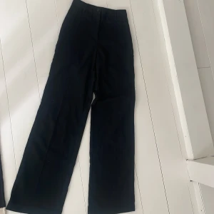 Svarta kostymbyxor från Monki - Svarta kostymbyxor från Monki med raka, vida ben och hög midja. Klassisk look med hällor för bälte och diskret knappstängning. Perfekta för en clean och stilren outfit. Materialet känns mjukt.