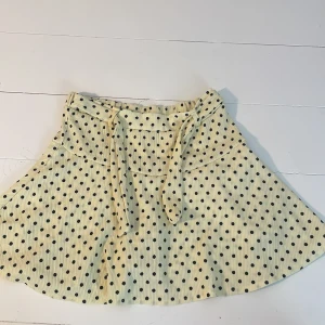 Prickig kortkjol från Zara med knyt - Supersöt kortkjol från Zara i ljusgul nyans med svarta prickar. Kjolen har knytband i midjan och är tillverkad i ett lätt, krinklat tyg som ger en skön känsla. Perfekt för dig som gillar lekfulla och trendiga plagg med retrovibbar.