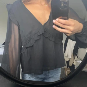 Svart blus med volang bikbok - Snygg svart blus från Bikbok med djup v-ringning och volangdetaljer framtill. Blusen har långa, transparenta ärmar och axelvolanger som ger en trendig look. Perfekt att styla till både kjol och jeans.