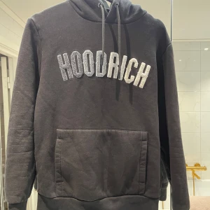 Svart hoodie från Hoodrich XS - Svart hoodie från Hoodrich i storlek XS med stor broderad logga framtill. Tröjan har huva med snörning och en stor känguruficka. Perfekt för chill dagar och streetstyle. Mjuk och skön bomullsblandning, långärmad modell.