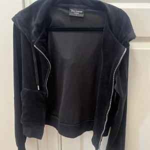 Juicy Couture svart kofta S - Svart velourkofta från Juicy Couture med huva, dragkedja med J-hänge och känguruficka framtill. Klassisk hoodie-stil med långa ärmar och ribbade muddar. Perfekt för en chill och trendig look.