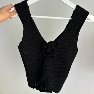 Svart virkad linnetopp med blomdetalj - Säljer en svart virkad linnetopp med v-ringning och en stor virkad blomma framtill. Toppen är ärmlös och har en lite croppad passform. Superfin till sommaren! XS/S