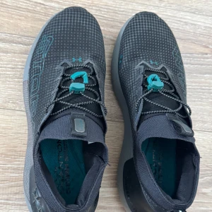 Svarta Under Armour sneakers HOVR Storm - Snygga svarta sneakers från Under Armour med HOVR-sula och Storm-detaljer. Ovandelen har rutigt mönster och turkosa accenter på snörningen. Skorna har elastisk mesh, reflekterande detaljer och platt sula för extra komfort. Perfekta för träning eller vardag.