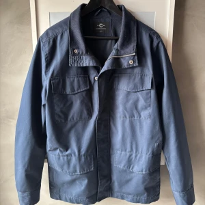 Mörkblå jacka från Selected Homme - Snygg mörkblå jacka från Selected Homme med fyra stora fickor framtill, hög krage och dragkedja med tryckknappar. Jackan har en clean och stilren look, perfekt för dig som gillar minimalistisk stil. Materialet är bomull och polyester.