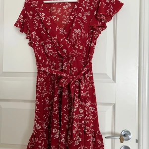 Röd blommig omlottklänning med volang - Supersöt röd kortklänning med vitt blommönster, omlottmodell och volangdetaljer på ärmarna. Klänningen har knytband i midjan och v-ringning. Perfekt för sommarens alla tillfällen!