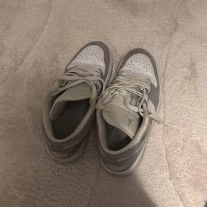 Nike Air Jordan 1 Low vit/grå - Snygga Nike Air Jordan 1 Low sneakers i vitt och ljusgrått skinn och mocka. Klassisk siluett med perforerad tå, snörning och ikoniska Jordan-loggor på plös och häl. Perfekta för dig som gillar stilrena och sportiga sneakers.