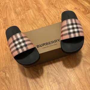 Burberry sandaler med klassiskt rutmönster - Snygga sandaler från Burberry med den ikoniska beige, svarta och röda rutan över foten. Svart sula med präglad logga och bekväm passform. Perfekta för sommaren och ger en lyxig touch till din outfit.