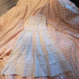 Vit somrig klänning  - Vit kortklänning med tunna axelband och blommor på tyget. Klänningen har volangdetaljer, snörning framtilll. Har använts 2 gånger.