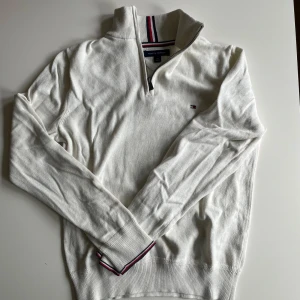 Vit half zip tröja Tommy Hilfiger - Vit långärmad tröja från Tommy Hilfiger med half zip och klassiska ränder i kragen och vid ärmslut. Tröjan är i mjuk bomull och har en liten broderad logga på bröstet. Perfekt för en clean och stilren look. Säljer för 200kr