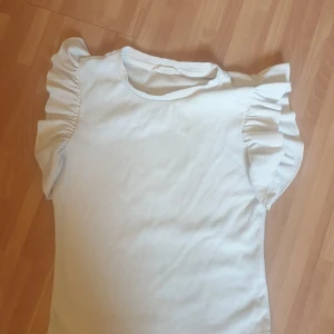 Vit volangtopp från SHEIN - Supersöt vit topp från SHEIN med ribbad struktur och volangärmar som ger en extra touch. Perfekt för dig som gillar en enkel men ändå trendig stil. Toppen är kortärmad och har en rund halsringning.