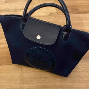 Svart handväska från Longchamp - Snygg svart handväska från Longchamp med präglad logga framtill. Väskan har två handtag, lock med silverfärgad knapp och dragkedja upptill. Tillverkad i en kombination av skinn och syntetmaterial, perfekt för dig som gillar stilrena accessoarer.