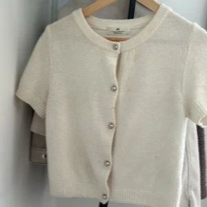 Beige kortärmad kofta från H&M - Superfin beige kofta från H&M med korta ärmar och rund hals. Koftan har silvriga knappar framtill och är stickad i ett mjukt material. Perfekt att slänga över en topp för en chill och stilren look. Tröjan är i storlek 158/164, 12-14y och passar till alla väder💕💐